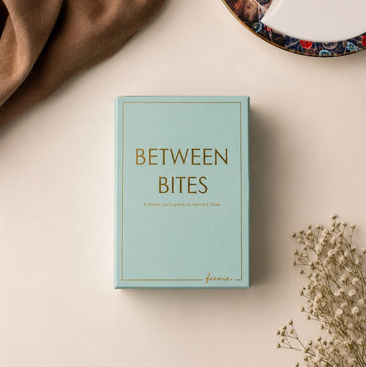 A mint green card deck  game titled 'BETWEEN BITES' with gold lettering, styled in a flat lay with a brown fabric corner, a decorative plate edge, and dried flowers on a light beige background. New Dinner Table card game by Feenies Table.
